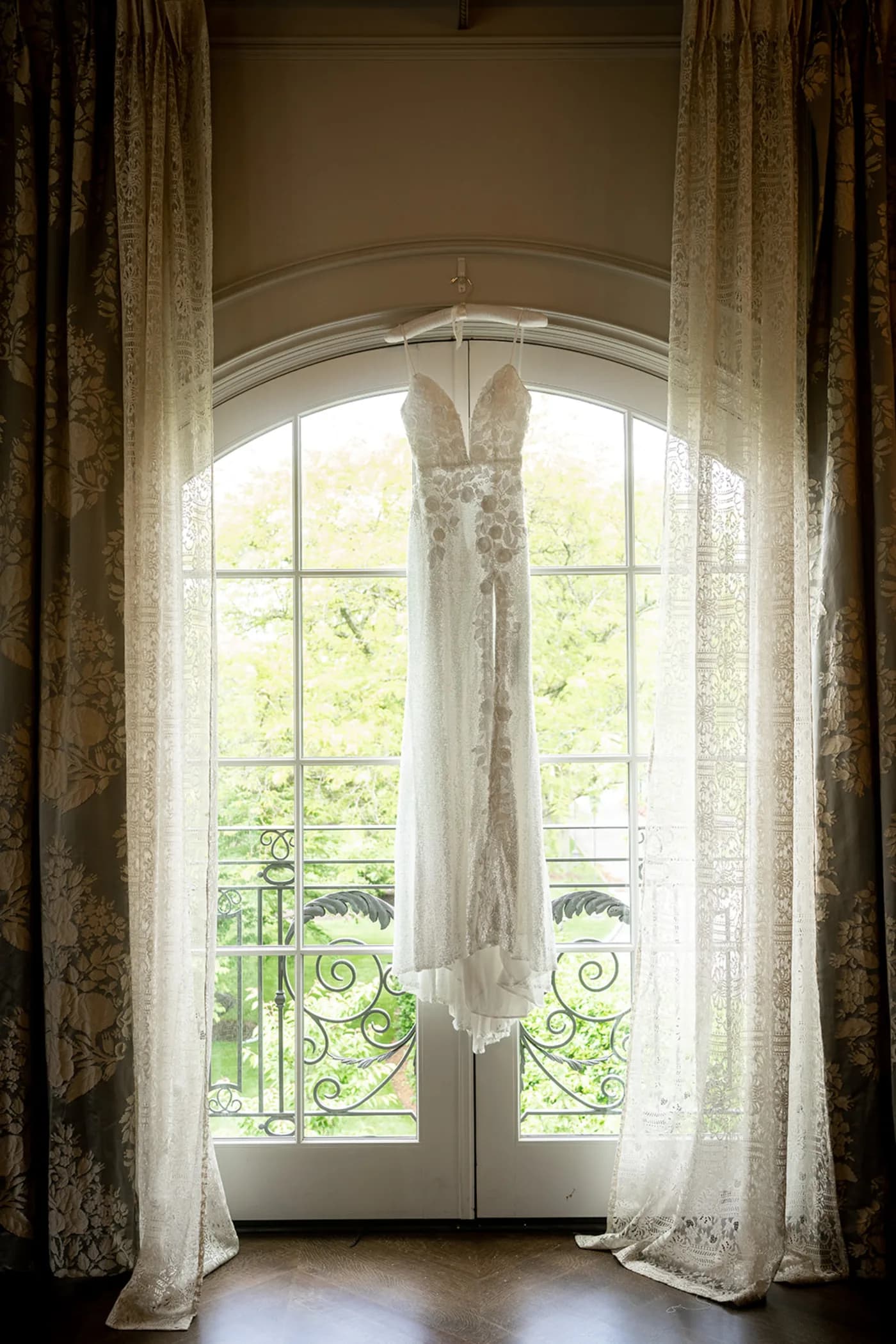 The wedding dress on a hanger in a tall arched window, framed by sheer curtains.